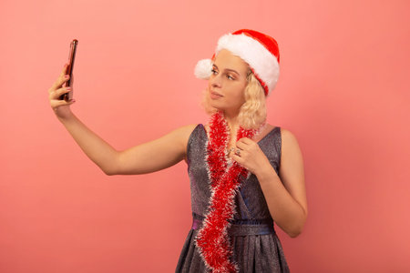 Young pretty woman taking selfie and wearing Santa Claus hat, silver sparkly glitter dress with red tinsel on a pink background. Girl taking photo and celebrating New Year.の写真素材