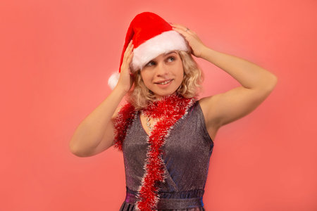 Happy cheerful smiling blonde girl in Santa Claus hat, silver sparkly dress with red tinsel looking up on pink background. New Year party and Christmas celebration concept.の写真素材