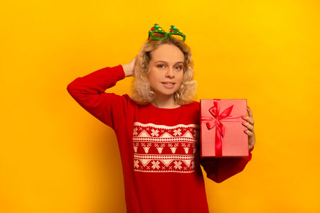 Happy young blonde woman holding pink present and looking at camera on a yellow background. New year concept. Smiling girl wearing red sweater and funny Christmas tree glasses.の写真素材