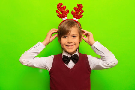 Happy smiling cute boy in Christmas reindeer antlers headwear, costume with black bow tie looking at camera on colorful green background. New Year and Xmas celebration concept.の写真素材
