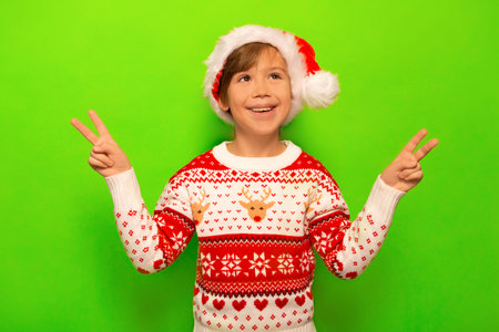 Happy smiling little boy in Santa Claus hat, Christmas sweater with deer and snowflakes pattern showing peace gesture or v sign with two fingers and looking up on green background.の写真素材