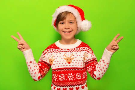 Happy smiling little child boy in Santa Claus hat, Christmas sweater with deer and snowflakes pattern showing peace gesture or v sign and looking at camera on a green background.の写真素材