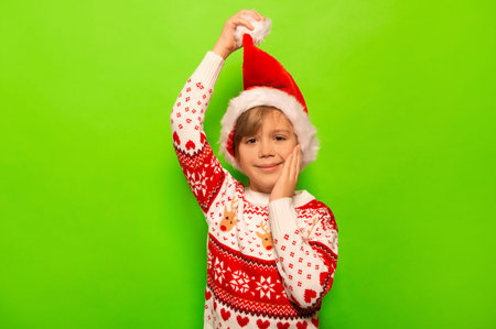 Happy smiling little boy raising Santa Claus hat, wearing Christmas sweater with winter pattern, keeping hand on cheek and looking at camera on a colorful green background.の写真素材