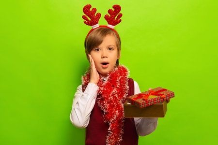 Surprised shocked cute boy in red Christmas reindeer antlers headwear with tinsel holding open present and looking at camera on green background.の写真素材