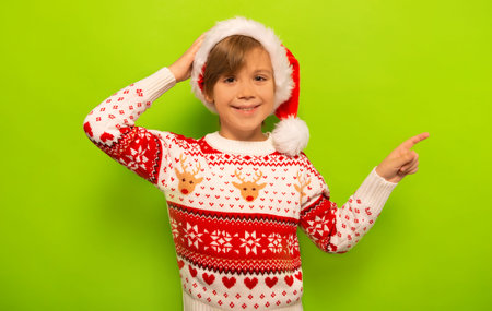 Happy smiling little boy in Santa Claus hat and Christmas sweater with winter pattern ornament pointing index finger to right side and looking at camera on a green background.の写真素材