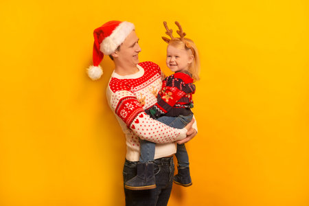 Happy smiling family father and daughter wearing Christmas sweaters, Santa Claus hat, reindeer antlers and laughing on yellow background. Man and little girl celebrating New Year.の写真素材