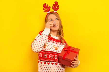 Happy dreaming young woman in Xmas reindeer antlers headwear, sweater with winter pattern holding present gift, Christmas ball instead of nose and looking up on yellow background.の写真素材