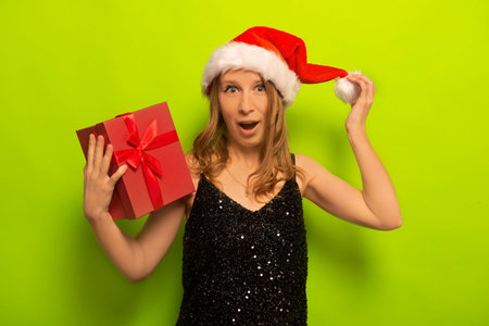 Surprised happy young woman in Santa Claus hat holding pink present gift box, wearing sparkly black dress and looking at camera on a green background. New Year and Xmas concept.の写真素材