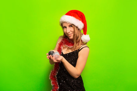 Happy smiling cheerful woman in Santa Clause hat holding Christmas balls, wearing sparkly black dress with red tinsel and looking at camera on a colorful green background.の写真素材