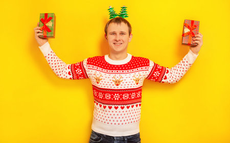 Happy smiling young man holding two wrapped presents gifts, wearing Christmas sweater with winter pattern, Xmas tree head wear and looking at camera on a yellow background.の写真素材