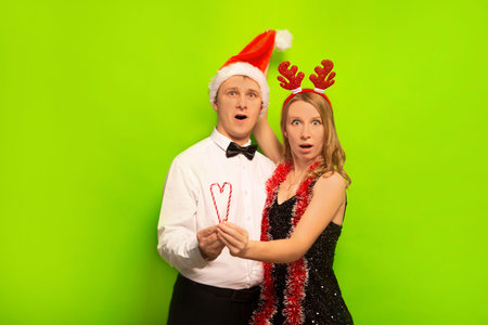 Surprised shocked young people husband and wife or friends wearing Santa Claus hat, reindeer antlers, looking at camera and holding Christmas lollipop candy on green background.の写真素材