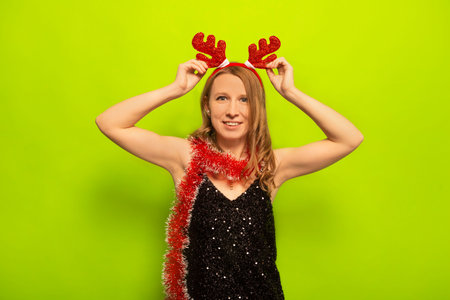 Happy smiling young woman in sparkly black dress with tinsel holding reindeer antlers headwear and looking at camera on colorful green background. New Year and Christmas concept.の写真素材