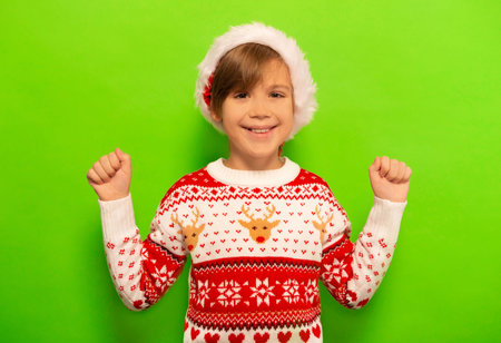 Happy smiling little kid boy in Christmas sweater and Santa Claus hat with raised hands or raised fists celebrating success, having fun and looking at camera on green background.の写真素材