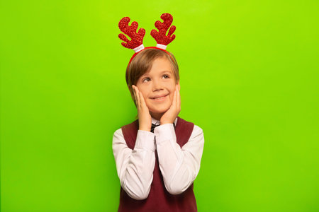 Happy little child boy in red Christmas reindeer antlers, white shirt with vest holding hands on cheeks or keeping hands on face and looking up. Kid dreaming on green background.の写真素材