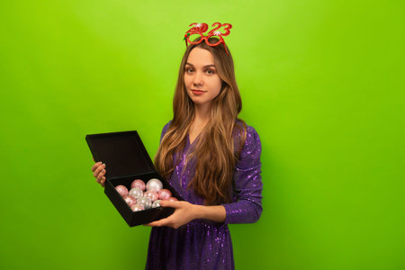 Cheerful young woman wearing party glasses 2023 with purple dress, holding black box with Christmas balls and looking at camera on green background. New Year 2023 concept.の写真素材