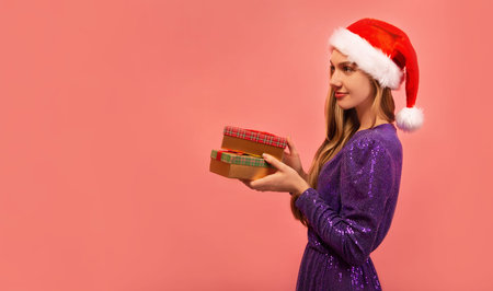 Happy cheerful young woman in Santa Claus hat and sparkly purple dress holding wrapped presents gifts on pink background with copy space. Happy New Year and Christmas celebration.の写真素材