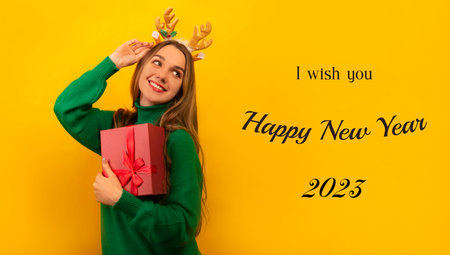 Happy smiling girl in Christmas reindeer antlers and sweater holding pink present gift box and looking up on a yellow background with text words I wish you Happy New Year 2023.の写真素材