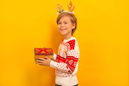 Happy smiling little child boy holding wrapped Christmas presents gifts, wearing winter Xmas sweater with reindeer antlers headwear and looking at camera on yellow background.の写真素材