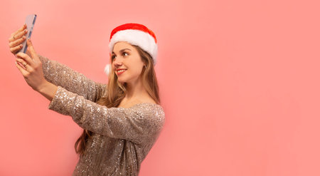 Happy smiling young woman in glitter dress with Santa Claus hat taking selfie photo on smartphone on pink background with copy space. Woman doing selfie shot on mobile phone.の写真素材