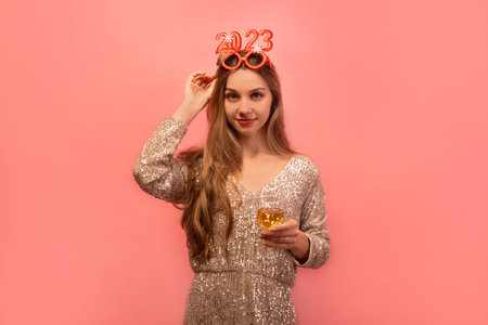 Young cheerful woman in sparkly glitter dress and party glasses celebrating New Year 2023, holding champagne glass or white wine glass and looking at camera on pink background.の写真素材