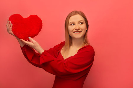 Happy smiling cheerful blonde girl holding red heart toy, wearing red dress with sleeves and looking up isolated on pink background. Happy St Valentine's Day celebration concept.の写真素材