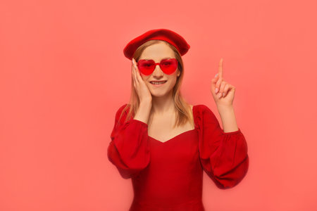 Happy smiling stylish girl pointing index finger up or holding finger up and keeping hand on face isolated on pink background. Woman in red dress, beret and heart shaped glasses.の写真素材