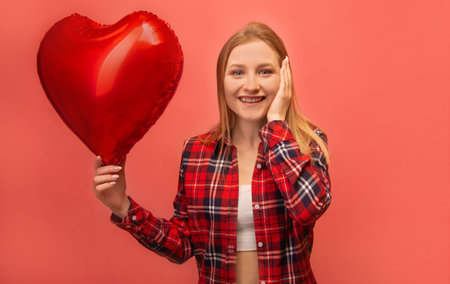 Surprised shocked happy smiling girl showing red heart shaped air balloon, holding hand on face and looking at camera on pink background. Dreamy girl in red checkered plaid shirt.の写真素材