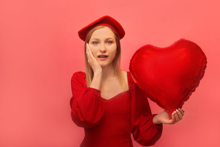 Stylish surprised shocked girl celebrating Valentines Day. Amazed woman in red dress, beret holding hand on cheek or face and showing heart shaped air balloon on pink background.の写真素材