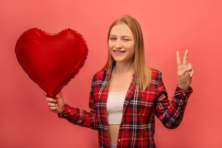 Happy winking girl showing peace gesture sign with two fingers and holding heart shaped air balloon. Smiling woman in casual red checkered plaid shirt isolated on pink background.の写真素材