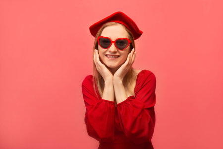 Happy surprised stylish young woman holding hands on face or keeping hands on cheeks, wearing red dress, beret with heart glasses and looking at camera isolated on pink background.の写真素材