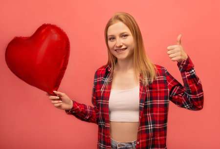 Happy girl showing thumb up, holding red heart shaped air balloon and looking at camera. Smiling woman in red checkered plaid shirt with white top isolated on pink background.の写真素材