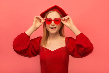Happy winking stylish woman in red dress with beret and heart shaped glasses showing tip of tongue, closing one eye and looking at camera isolated on pink background.の写真素材