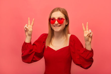 Happy smiling girl showing peace gesture v sign with four fingers and looking aside isolated on pink background. Stylish young woman in red dress and trendy heart shaped glasses.の写真素材