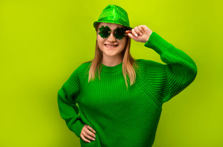 St Patrick's Day celebration concept: happy smiling blonde girl wearing green sweater with shiny party hat and holding clover glasses isolated on green background.の写真素材