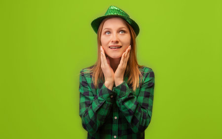 Surprised shocked happy young woman in green party hat and checkered plaid shirt holding hands on face and looking up isolated on green background. St Patrick's Day concept.の写真素材