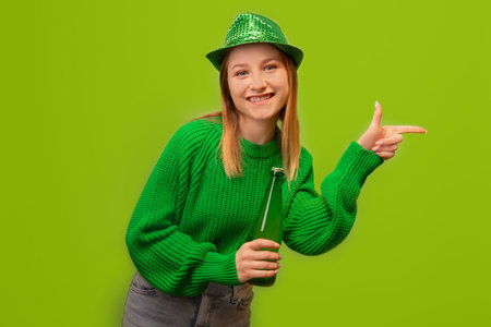 St Patrick's Day concept: happy smiling girl pointing index finger aside, showing offer, wearing green sweater with party hat and holding green beer bottle on green background.の写真素材