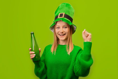 Happy smiling girl in St Patricks Day leprechaun green hat holding green beer bottle, rejoicing, saying yes, celebrating victory and keeping fist pump gesture on green background.の写真素材