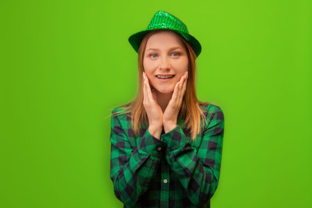 Surprised shocked happy young woman in green party hat and checkered plaid shirt holding hands on face and looking at camera isolated on green background.の写真素材