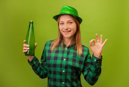 Cheerful positive girl in party green hat and checkered plaid shirt holding green beer bottle and showing ok gesture sign isolated on green background. St Patrick's Day concept.の写真素材