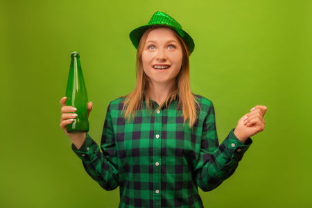 Surprised shocked excited woman in green party hat and checkered plaid shirt holding green beer bottle and looking up isolated on green background. St Patrick's Day celebration.の写真素材
