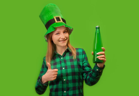 Happy smiling girl in green leprechaun hat and checkered plaid shirt recommending green beer bottle, showing thumb up or holding finger up on green background. St. Patrick's Day.の写真素材