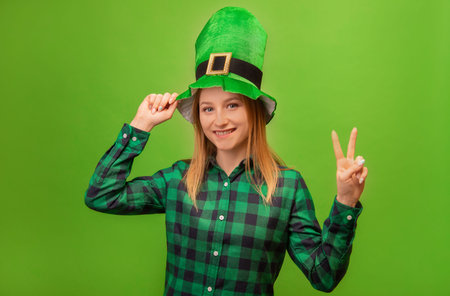 St Patricks Day leprechaun girl in green festive irish hat and checkered plaid shirt showing peace gesture with two fingers and looking at camera isolated on green background.の写真素材