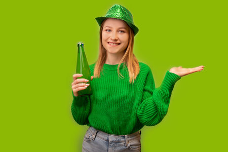 Happy smiling positive girl in green sweater with party hat holding green beer bottle and pointing hands arms aside at empty space on green background. St. Patrick's Day concept.の写真素材