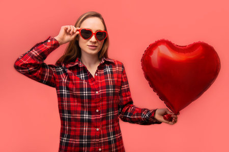 Stylish young woman holding red heart shaped air balloon and wearing casual checkered plaid shirt with party heart glasses isolated on pink background.の写真素材