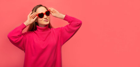 Stylish happy cheerful girl in warm pink dress with heart shaped glasses looking up on pink background with copy space. St Valentines Day,Women's Day or lifestyle concept. Banner.の写真素材