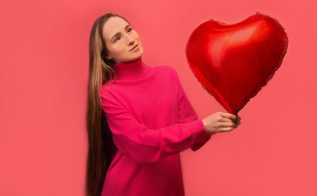 Dreaming stylish young woman holding red heart shaped air balloon and looking up isolated on pink background. Saint Valentines Day, happy birthday, Womens Day or love concept.の写真素材