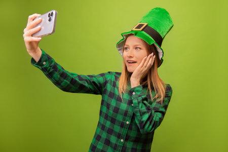 Happy shocked surprised girl in green leprechaun hat and checkered plaid shirt taking selfie on smartphone and holding hand on face on green background. St Patricks day concept.の写真素材