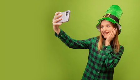 Happy smiling girl in green leprechaun hat and checkered plaid shirt taking selfie on smartphone and holding hand on face on green background with copy space. St. Patrick's day.の写真素材