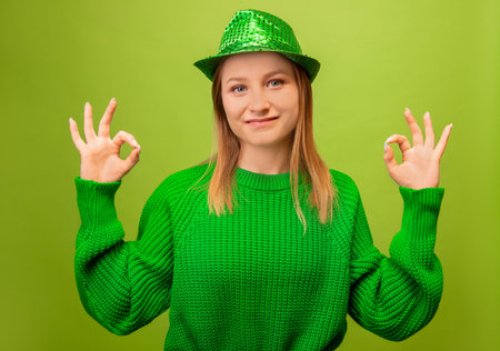 Cheerful smiling young blonde woman in green sweater with shiny hat gesture showing ok sign hand and looking at camera isolated on green background. St Patrick's Day concept.の写真素材