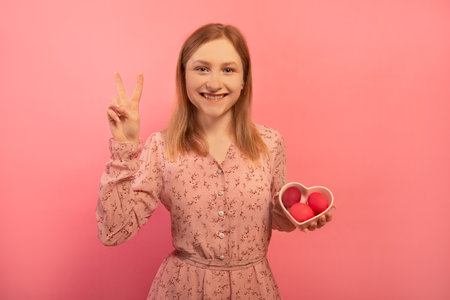 Happy smiling woman shows peace gesture v sign with two fingers hand and holds heart shaped plate dish with three dyed painted easter eggs isolated on pink background. Easter Day.の写真素材